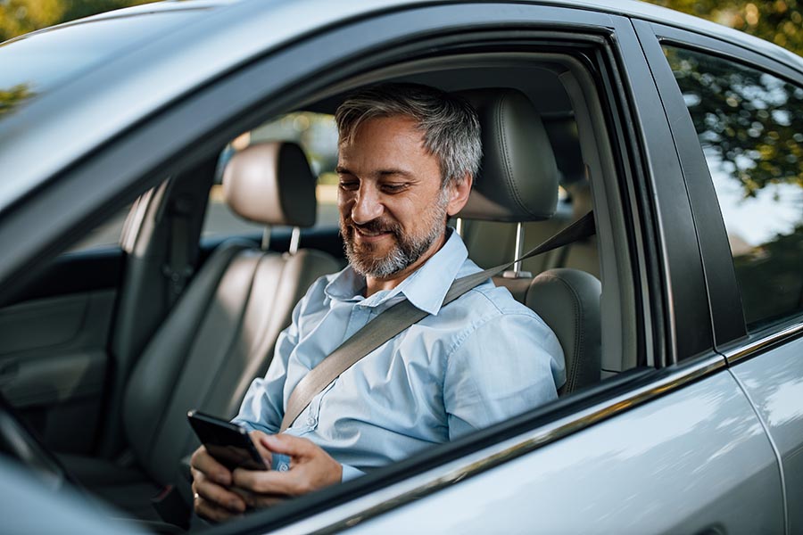 Client Center - Man in a Button Down Shirt With Graying Hair Smiles as He Uses a Smartphone in His Silver Car
