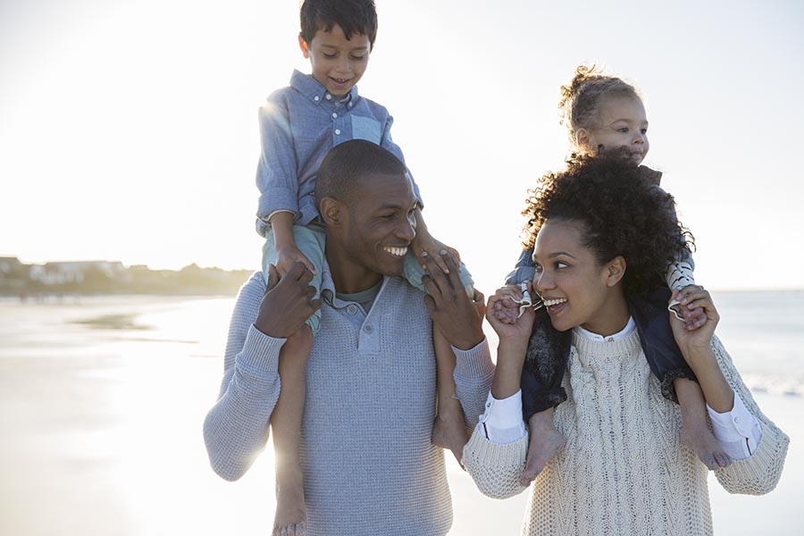 Personal Insurance - a Family of Four Strolls Along a Beach on a Cold Day, Son and Daughter Riding on Top of Their Parents' Shoulders, Everyone Smiling